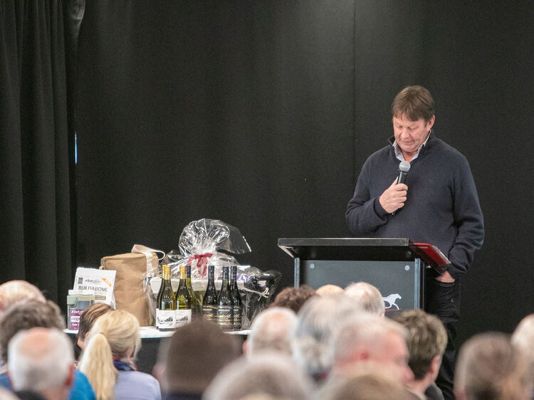 Speaker addresses a seated crowd at a harness racing event, with wine bottles and gift baskets on the table beside him