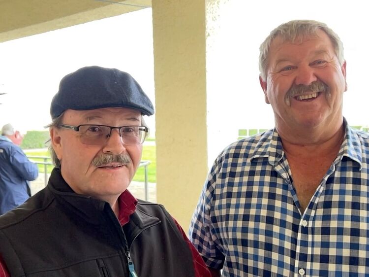 Two men stand together smiling outdoors, one in a cap and glasses, the other in a checkered shirt
