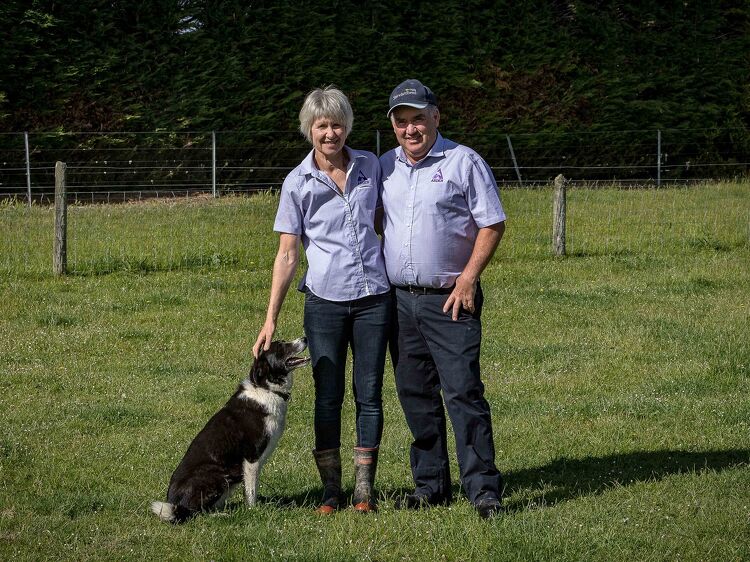 Couple standing in a grassy paddock with their dog, both wearing matching farm uniforms