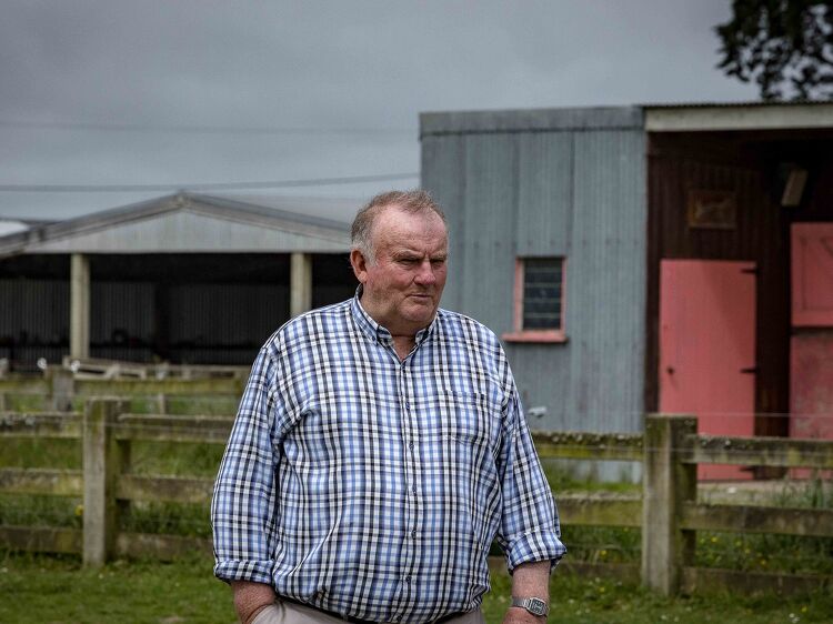 Older man in a checkered shirt stands outside near horse stables and fencing on a cloudy day